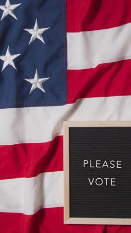 Vertical-Video-Of-Animated-Sign-Spelling-Out-Please-Vote-Lying-On-US-Stars-And-Stripes-Flag-For-American-Election-2024